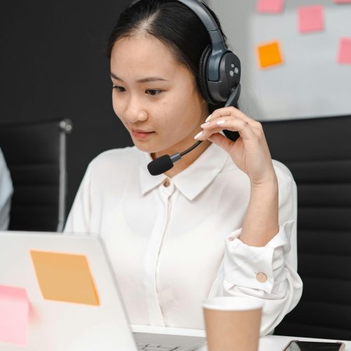 Asian woman using a laptop and headset for customer support in an office environment.